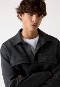 Camicia grigia scuro con bottoni, realizzata in tessuto morbido, con due tasche sul petto e una maglietta bianca a girocollo sotto. Dettagli in metallo argentato.
