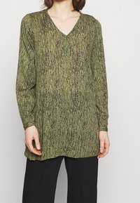 Groene tuniek met lange mouwen en een V-hals, voorzien van een gestructureerd zwart patroon dat doet denken aan verticale lijnen, gecombineerd met zwarte broeken.