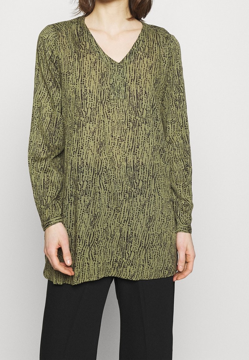 Groene tuniek met lange mouwen en een V-hals, voorzien van een gestructureerd zwart patroon dat doet denken aan verticale lijnen, gecombineerd met zwarte broeken.
