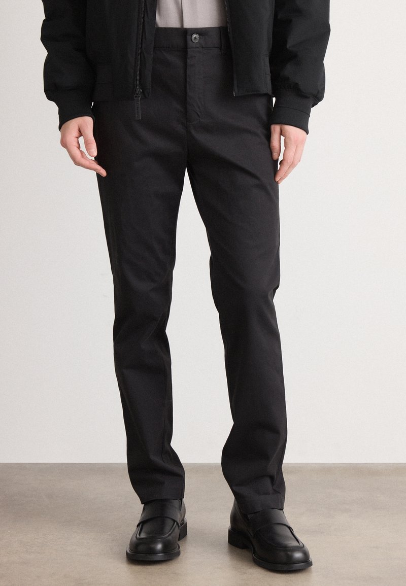 Homme portant un pantalon noir slim avec des mocassins en cuir noir, debout sur un sol uni sur fond blanc.