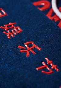 Caracteres japoneses en relieve de color rojo sobre una superficie de tela texturizada de color azul oscuro, dispuestos en diagonal con un enfoque variable.
