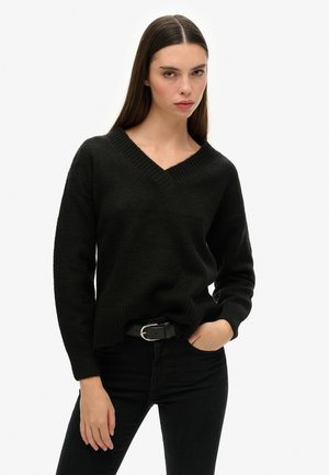 Suéter negro de punto con escote en V, mangas largas y un corte relajado. La textura es suave, combinado con jeans negros y un cinturón negro.