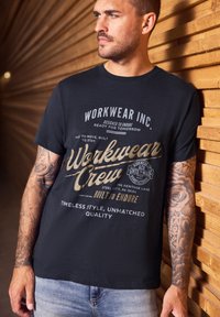 Muž s tetovanými rukama, oblečený v černém tričku s potiskem "Workwear Crew" a modrých džínách, opřený o dřevěnou stěnu s lamelami.