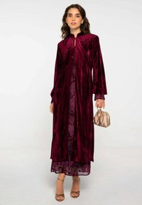 Robe en velours bordeaux à manches longues et col montant, superposée à une robe prune à motif floral. Les détails comprennent un petit sac à main et des talons à lanières.