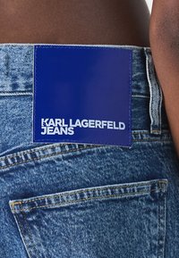 Denimske hlače z modrim umetnim usnjem na hrbtni strani, z belim napisom "KARL LAGERFELD JEANS". Vključuje detajl zadnjega žepa.