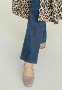 Zapatos Mary Jane brillantes de color plateado con una correa, usados con pantalones de mezclilla azul oscuro y un abrigo de estampado de leopardo. Textura suave y diseño de punta redonda.