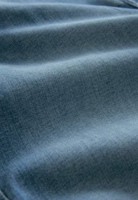 Tissu en denim d'une teinte bleue moyenne, présentant une texture douce et un motif de tissage diagonal visible. Le tissu est légèrement froissé.