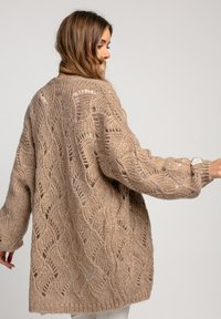 Beżowy sweter z dzianiny o luźnym kroju, z teksturowanym wzorem falistym, długimi rękawami i otwartym przodem. Miękki i przytulny materiał.