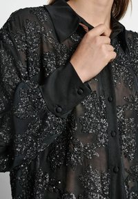 Must mustrits läikivast mustast materjalist blouse, millel on lilleline sädelusmuster, nööbiga eesmine osa, klassikaline krae ja nööbiga varrukate äär, mis loob tekstuurse ja kihilise välimuse.