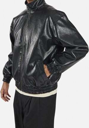 Hombre con chaqueta bomber de cuero negro brillante con puños y dobladillo elásticos, una mano en el bolsillo, combinada con pantalones negros y camiseta blanca.