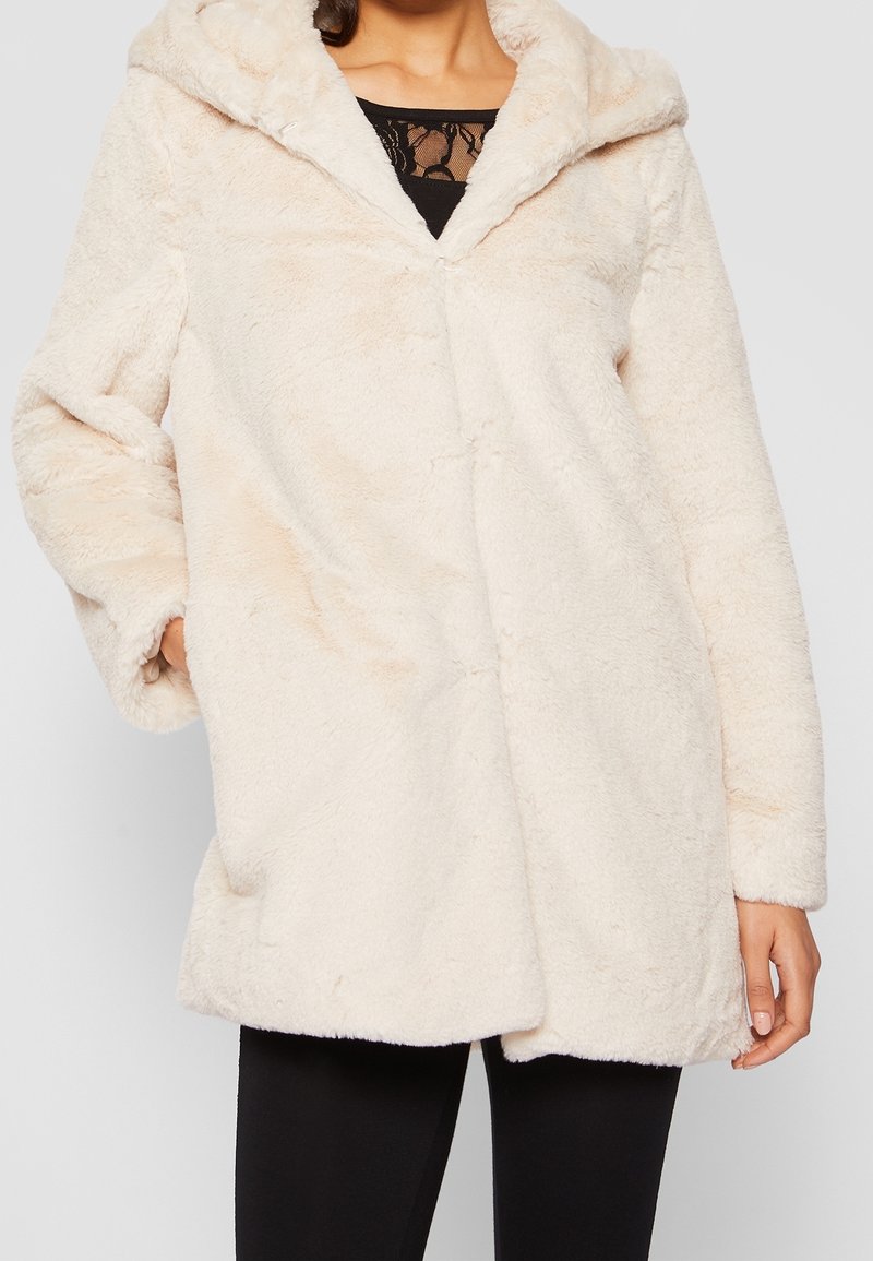 Vrouw in een crèmekleurige, gewatteerde faux fur jas, met een capuchon, over een zwart kant top en zwarte broeken, met één hand in de jaszak.