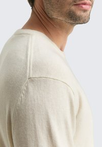 Mann in einem cremefarbenen Strickpullover, gezeigt von den Schultern bis zur Mitte des Halses, mit leichten Stoppeln im Gesicht.