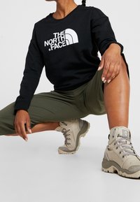 Felpa nera con logo bianco, pantaloni verde oliva e stivali da trekking beige con superficie texture e occhielli in metallo.