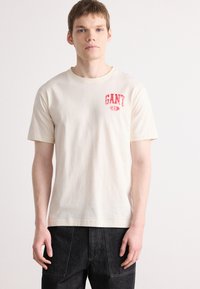 Krém színű, rövid ujjú pamut póló. Az elülső bal mellen vörös "GANT" logó található. Egyszerű, laza szabású design.