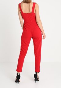 Tuta rossa con spalline larghe, vita aderente e gambe tapered. Realizzata in tessuto liscio. Indossata con scarpe con tacco alto nere. Dettaglio con zip sul retro.
