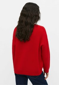 Personne aux longs cheveux ondulés et foncés portant un pull en laine rouge et un jean bleu foncé, debout contre un fond blanc uni, de dos.