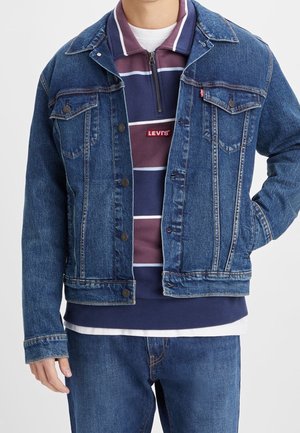 Personne portant une veste en jean bleue et un jean par-dessus un pull Levi's à fermeture éclair quart, rayé bordeaux et marine, avec un sous-maillot blanc.