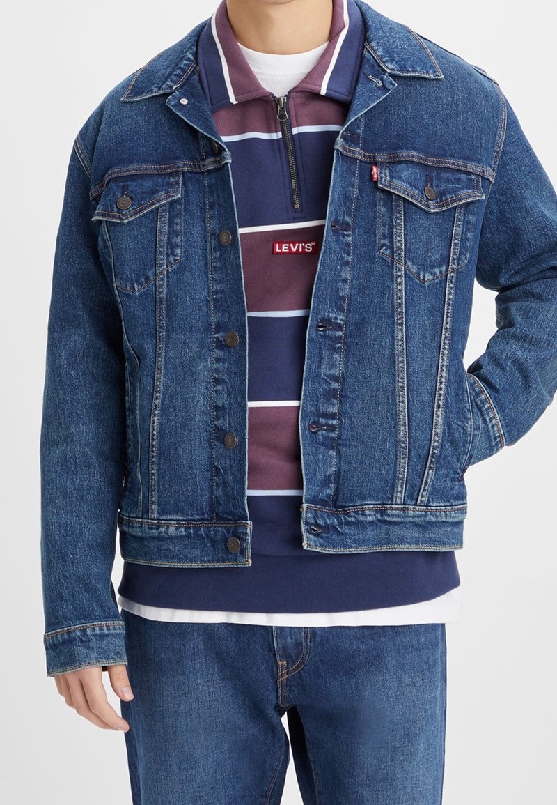 Personne portant une veste en jean bleue et un jean par-dessus un pull Levi's à fermeture éclair quart, rayé bordeaux et marine, avec un sous-maillot blanc.