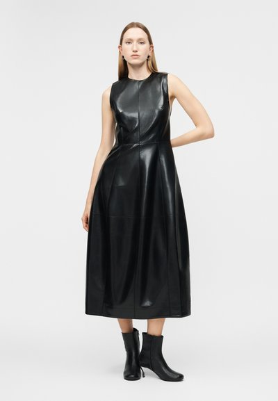 Γυναίκα που φοράει μαύρο δερμάτινο μίντι φόρεμα χωρίς μανίκια και μαύρα μποτάκια μέχρι τον αστράγαλο, στέκεται με το ένα χέρι στη μέση της μπροστά σε λευκό φόντο.