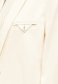 Μπεζ σακάκι με ένα προσεκτικά διπλωμένο λευκό μαντηλάκι στην τσέπη στο αριστερό μέρος του στήθους.