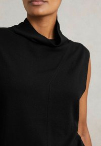 Mustamuguline lühikeste varrukateta topp, millel on volditud kõrge krae. Valmistatud pehmest kangast, esiküljel on diagonaalne õmbluse detail. Lihtne, minimalistlik disain.