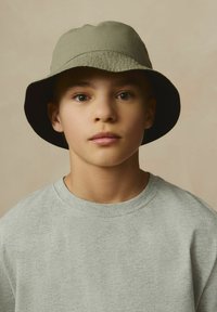 Grijze katoenen sweatshirt gedragen met een donkergerande olijfgroene vissershoed. De hoed heeft een brede, flexibele rand en een gestructureerde stof.