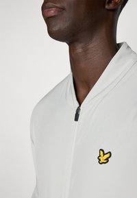 Light grey zip-up jacket featuring a hidden zipper, stand collar, and a yellow bird logo on the left side near the neck.