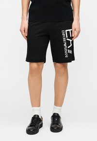 Μαύρα βαμβακερά σορτς με ίσια γραμμή, με λευκό κάθετο λογότυπο στην αριστερή πλευρά. Φοριούνται με μαύρα αθλητικά παπούτσια και λευκές κάλτσες.