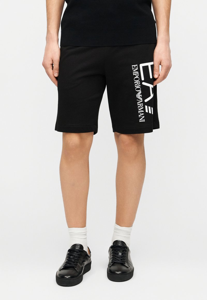 Μαύρα βαμβακερά σορτς με ίσια γραμμή, με λευκό κάθετο λογότυπο στην αριστερή πλευρά. Φοριούνται με μαύρα αθλητικά παπούτσια και λευκές κάλτσες.