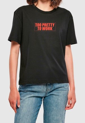 Persona con camiseta negra con texto rojo "DEMASIADO BONITA PARA TRABAJAR" combinada con vaqueros azul claro sobre un fondo liso.