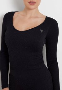 Schwarzes langärmeliges Top aus geripptem Stoff mit Rundhalsausschnitt. Verfügt über eine dezente glitzernde Textur und ein kleines dreieckiges Logo auf der linken Seite.