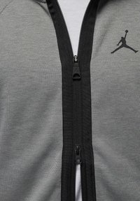 Giacca sportiva grigia con zip e rifiniture nere. Presenta un logo nero e una texture liscia, progettata per il comfort e la mobilità.