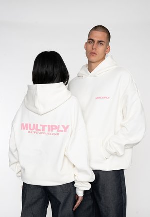 White hoodies with pink "MULTIPLY" logo on back and front, featuring a relaxed fit and kangaroo pocket, paired with dark jeans.