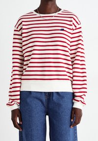 Sweat-shirt rayé rouge et blanc avec un col rond côtelé, des manches longues et un petit logo bleu sur la poitrine. Associé à un jean en denim bleu.