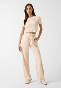 Heller beiger, verkürzter T-Shirt mit "GUESS" aus glänzenden Nieten, kombiniert mit passenden, lockeren Hosen mit Kordelzug in der Taille. Sanfte Stofftextur.