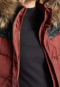 Personne portant un manteau d’hiver bordeaux avec une capuche en fausse fourrure et un haut noir en dessous, manteau partiellement ouvert, haut du torse visible.