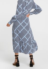 Maxiklänning i svart och blå geometrisk mönster, långärmad, volangkant och åtsittande midja; bärs med svarta remsandaler.