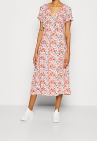 Abito midi floreale con maniche corte a volant, scollatura a V profonda, prevalentemente bianco con motivi floreali rossi, blu e rosa, abbinato a sneakers bianche.
