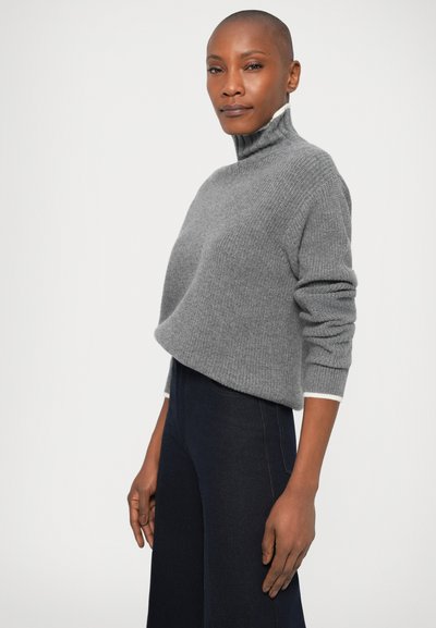 Pull en tricot gris avec un col haut, présentant un tissu texturé et un accent blanc aux poignets, associé à un pantalon en denim foncé.