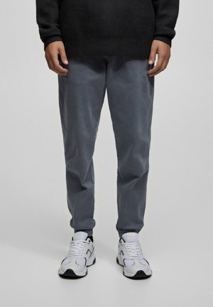 Personne portant un pantalon gris foncé, des baskets blanches avec des détails noirs et un pull noir, debout sur un fond clair uni.