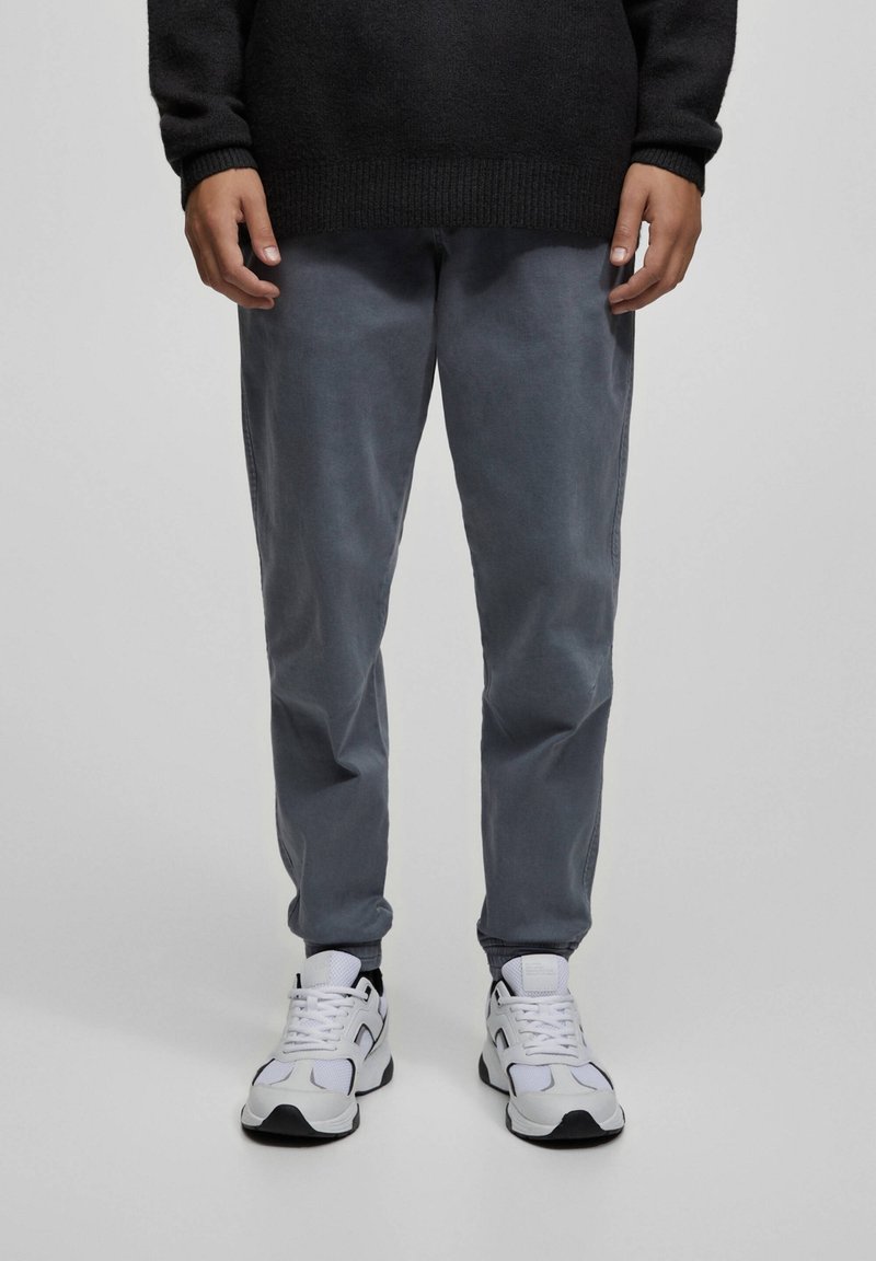 Personne portant un pantalon gris foncé, des baskets blanches avec des détails noirs et un pull noir, debout sur un fond clair uni.