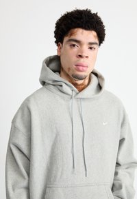 Pelēks kapucēnš, kas izgatavots no mīksta materiāla, ar priekšējo kabatu, auklām un mazu baltu Nike logotipu uz krūšu daļas.