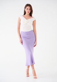 Top bianco aderente con design attorcigliato e accento di rosa; gonna di satin lilla che scende fino al polpaccio; la modella indossa tacchi nudi con cinturini.