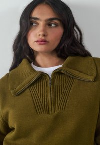 Olivgrüner gestrickter Pullover mit Reißverschlusskragen, gerippter Textur am Kragen und an der Vorderseite sowie einem weichen Finish.