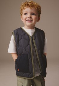 Vestă matlasată în gri închis cu margini verde măsliniu, cu fermoar frontal, două buzunare laterale și o textură netedă, ușoară. Poartă peste o cămașă albă.