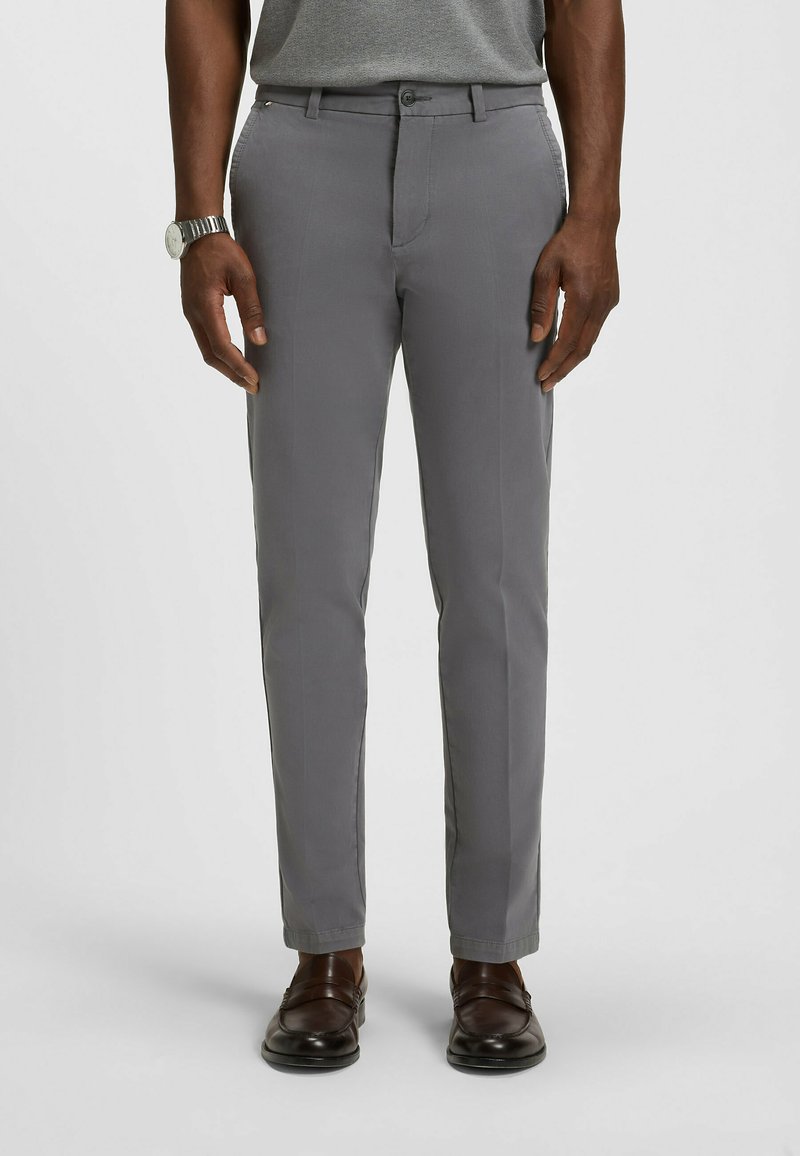 Mężczyzna ubrany w szare, dopasowane spodnie, brązowe skórzane loafersy, szarą koszulę i srebrny zegarek na rękę, stojący na tle gładkiej powierzchni.