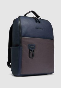 Mochila moderna rectangular en azul marino y marrón con asa superior, bolsillo frontal con cremallera y mosquetón adjunto sobre fondo gris.