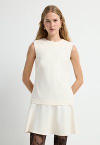 Mouwloze crèmekleurige jurk met een gestructureerd lijfje en een uitlopende rok. Gladde textuur met een subtiele naadaccent bij de taille. Zwarte getailleerde maillot.