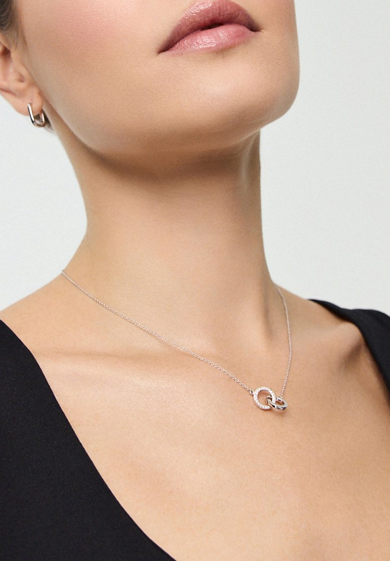 Widać szyję i podbródek kobiety, która nosi delikatny srebrny naszyjnik z przeplatającymi się pierścionkami, pasujący do jej gładkiego czarnego topu.