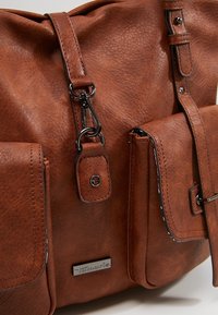 Sac en cuir marron avec deux poches avant, détails de fermoirs métalliques, une bandoulière amovible et une étiquette métallique de la marque "Tamaris".